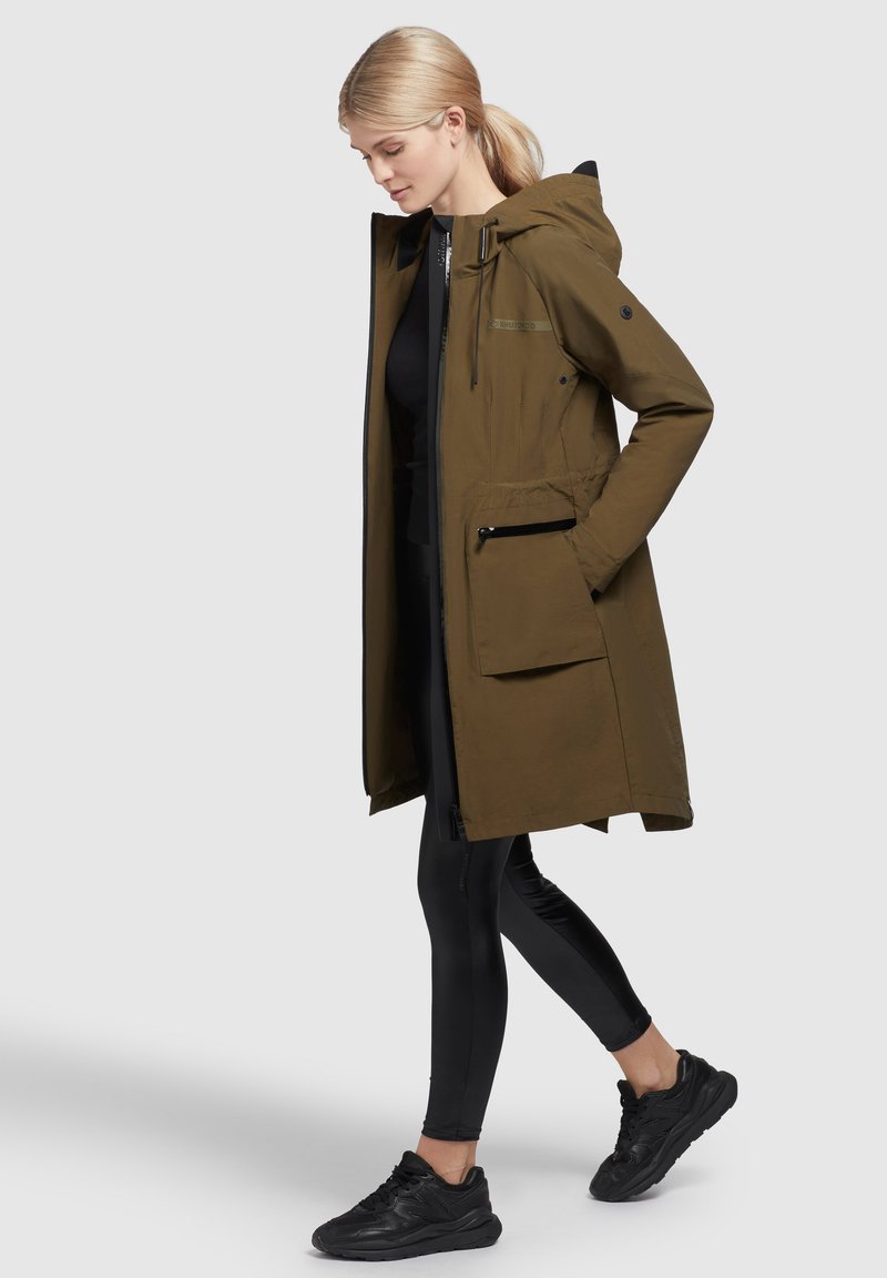 Olivenfarbener wasserdichter Parka mit Kapuze, Reißverschluss vorne und Seitentaschen; getragen über schwarzen Leggings und schwarzen Sneakers.