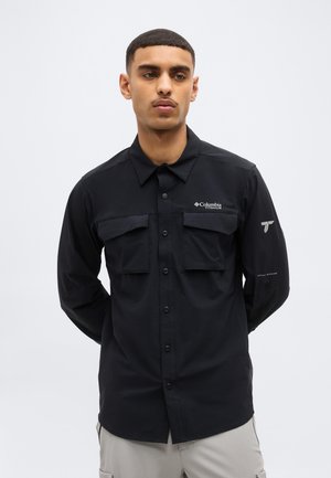 Rövid hajú fiatal férfi fekete, hosszú ujjú, zsebes, gombos ingben, amelyen logók találhatók a mellkason és az ujjon.