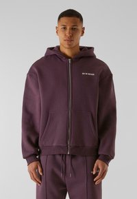 Hoodie zippé violet en tissu doux, avec une poche kangourou et un petit logo sur la poitrine. Disponible avec un pantalon de survêtement assorti.