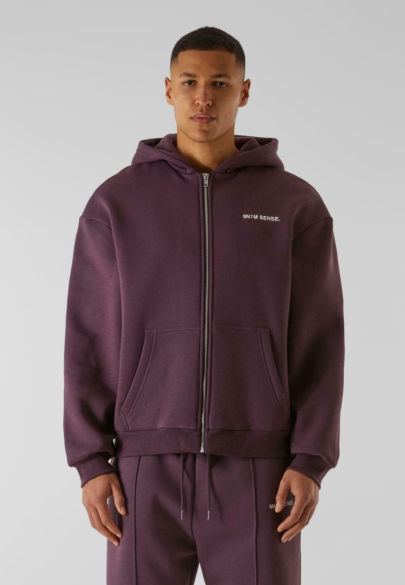 Hoodie zippé violet en tissu doux, avec une poche kangourou et un petit logo sur la poitrine. Disponible avec un pantalon de survêtement assorti.