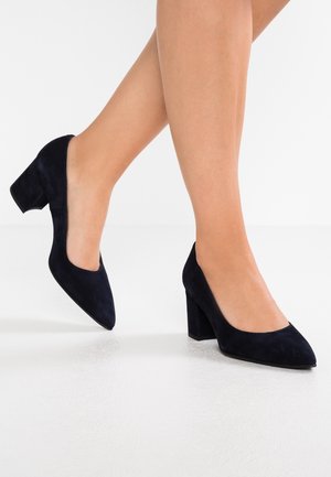 Jambes de femme portant des escarpins à talons blocs en daim noir à bouts pointus sur un fond de sol blanc.
