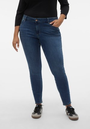 Persona que lleva vaqueros ajustados azul oscuro, camiseta de manga larga negra y zapatillas negras y blancas, de pie con una mano en el bolsillo.