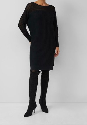 Femme portant une robe noire côtelée à manches longues avec des manches supérieures transparentes et des bottes noires à talons au-dessus du genou, debout contre un mur blanc.