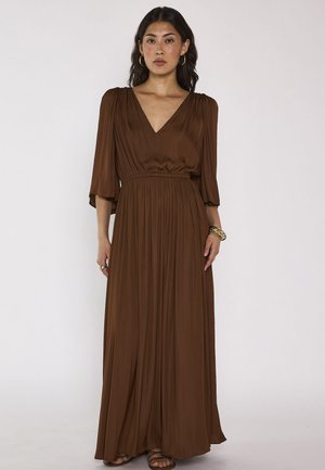 Donna che indossa un lungo abito plissettato marrone con maniche tre quarti e scollo a V, accessoriata con orecchini a cerchio dorati e braccialetti.