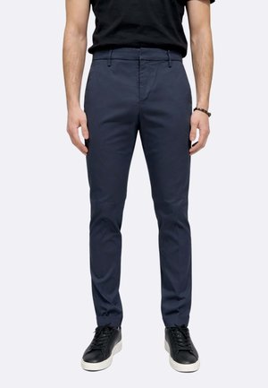 Uomo che indossa pantaloni slim fit blu navy, scarpe da ginnastica nere con suole bianche e una t-shirt nera, in piedi contro uno sfondo chiaro.
