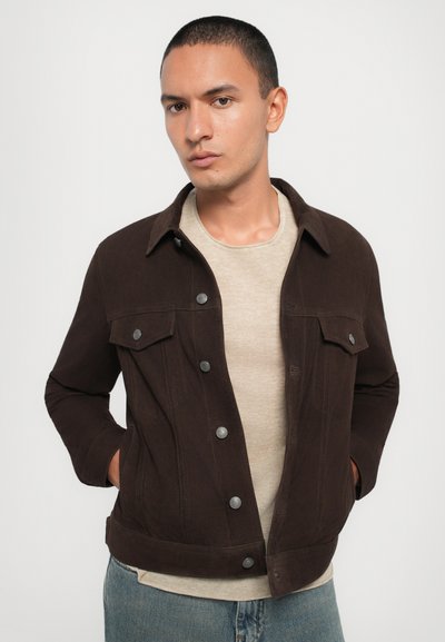 Veste en denim marron avec des boutons-pression argentés, deux poches poitrine et une coupe ajustée. Portée sur une chemise beige, stylée avec un jean bleu.