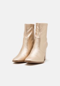 Bottines à bout pointu en or métallisé. Fabriquées en matériau synthétique lisse avec une fermeture éclair et un talon haut effilé. Texture douce.