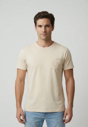 Hombre con cabello corto y oscuro, vistiendo una camiseta beige de manga corta con un bolsillo en el pecho y jeans azul claro, posando frente a un fondo liso.