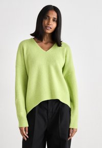 Gaiši zaļš V-veida kakla džemperis, izgatavots no mīksta auduma ar brīvu piegriezumu, ribotām piedurknēm un nedaudz garāku aizmugurējo apakšmalu, kombinēts ar melnām biksēm.