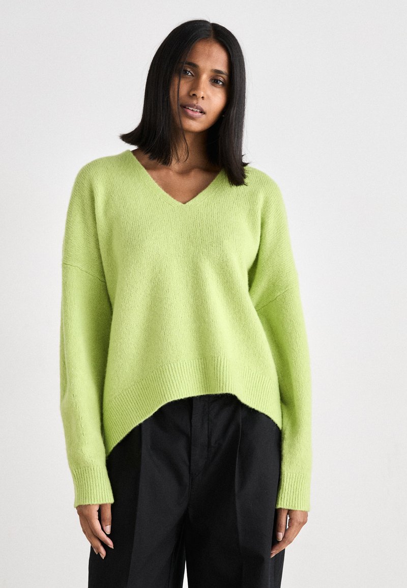 Gaiši zaļš V-veida kakla džemperis, izgatavots no mīksta auduma ar brīvu piegriezumu, ribotām piedurknēm un nedaudz garāku aizmugurējo apakšmalu, kombinēts ar melnām biksēm.