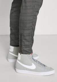 Ljusgrå högsneakers med vita accenter, placerade mot svarta och grå rutiga byxor med sidsdragkedjor. Texturerat mockamaterial.