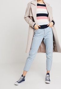 Jasnobeżowy płaszcz nałożony na sweter w paski w odcieniach różu, niebieskiego i białego. Niebieskie dżinsy i granatowe sneakersy. Prosty, nowoczesny design.