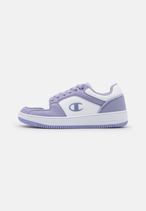 Zapatilla baja en blanco y lavanda con cordones, suela plana y un logotipo en el lateral, mostrada desde el perfil exterior.