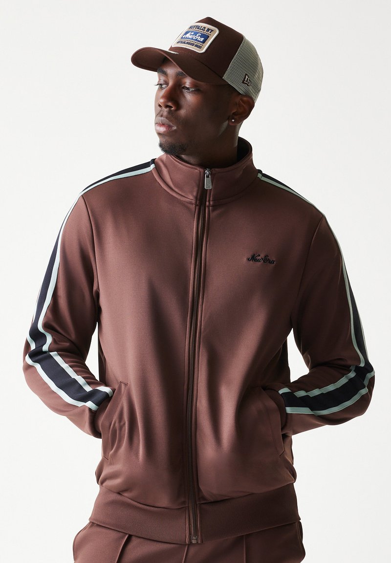 Καφέ ζακέτα με φερμουάρ,  ψεύτικο γιακά και πλαϊνές τσέπες. Διακοσμημένη με λευκές και ναυτικές ρίγες στα μανίκια. Συνδυάζεται με γκρι καπέλο από δίχτυ.