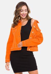 Orangenes Jeansjacke mit weißem Nähten, taillierter Schnitt und Taschen auf der Vorderseite, getragen über einem schwarzen gerippten Kleid. Silberne Druckknöpfe akzentuieren die Jacke.