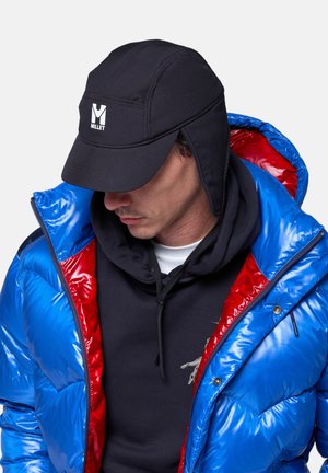Homme portant un chapeau en polaire noir avec des cache-oreilles, une doudoune brillante bleue et rouge, et un sweat à capuche noir, regardant vers le bas et sur le côté.