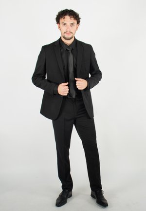 Costume noir avec un design ajusté, manches longues et revers en crans, associé à une cravate noire et des chaussures formelles, sur un fond uni.