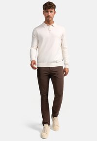 Polo de manga larga beige con collar y tres botones, combinado con pantalones slim-fit marrones y zapatillas de tonos claros.