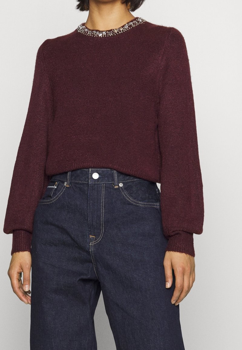 Viininpunainen neulepaita teksturoidulla pinnalla ja koristellulla pääntiellä, yhdistetty tummansinisiin korkeavyötäröisiin farkkuihin, joissa on kontrastitikkaukset.