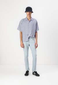 Camisa de manga corta abotonada en azul claro con un patrón geométrico, combinada con jeans desgastados azul claro y zapatillas negras.