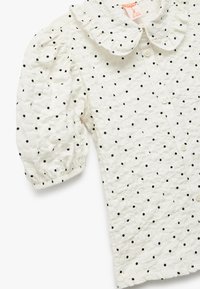 Blusa blanca de manga corta con collar rizado, que presenta lunares negros y mangas abullonadas recogidas. Cierre frontal con botones.