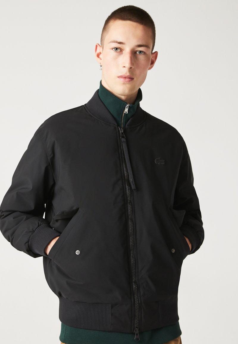 Jeune homme aux cheveux courts portant une veste bombardier noire zippée, les mains dans les poches, sur un fond blanc uni.