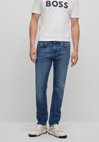 Man med blå slim-fit jeans, vita sneakers och en vit t-shirt med "BOSS" tryckt på bröstet, stående mot en enkel bakgrund.