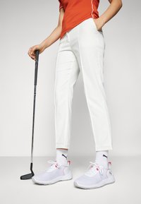 Weiße Golfhose, rotes kurzärmliges Hemd und leichte weiße Sneaker mit pinken Akzenten. Golfclub in der linken Hand.