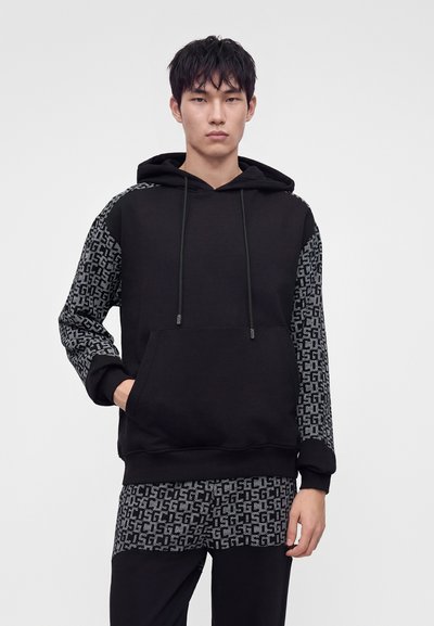 Jeune homme portant un sweat à capuche noir avec des manches grises à motifs et une chemise assortie à motifs en dessous, debout devant un fond uni.