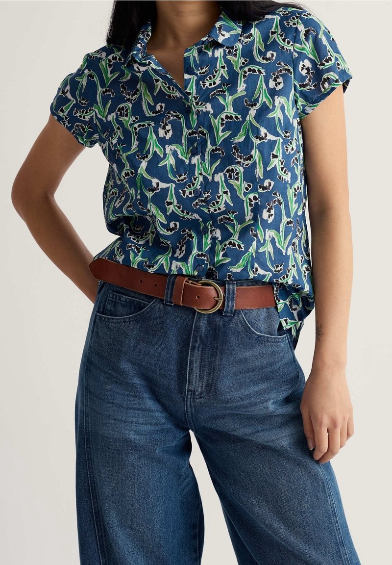 Femme portant une chemise fleurie bleue à manches courtes rentrée dans un jean bleu taille haute avec une ceinture marron, debout avec une main sur la hanche.