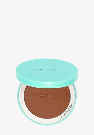 Bronzer-Compact mit einem türkisfarbenen Deckel und weißem Rand, mit eingeprägtem Markennamen. Das innere Puder hat einen warmen Braunton und eine strukturierte Oberfläche.