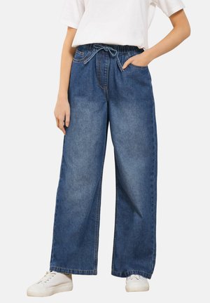 Persoon draagt bredere blauwe spijkerbroek met elastische taille en trekkoord, gecombineerd met witte sneakers en een effen wit T-shirt.