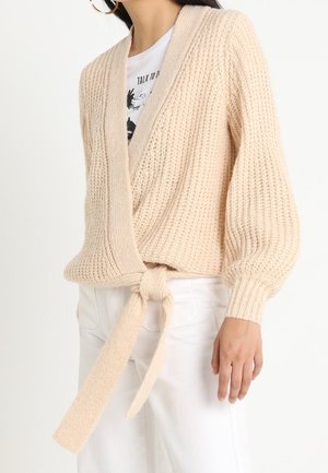 Femme portant un cardigan en maille beige à nouer à la taille, sur un t-shirt blanc graphique et un pantalon blanc, visible des épaules aux hanches.