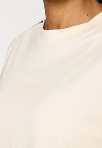 Osoba nosząca kremowy T-shirt z okrągłym dekoltem, z delikatnym wytłoczonym logo na piersi, ukazująca szyję i obszar ramion.