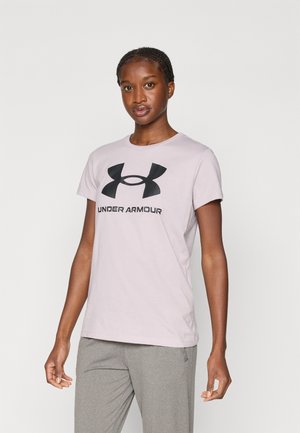 Kobieta stoi, mając na sobie jasno-różową koszulkę Under Armour z czarnym logo i szare spodnie na jednolitym jasnym tle.