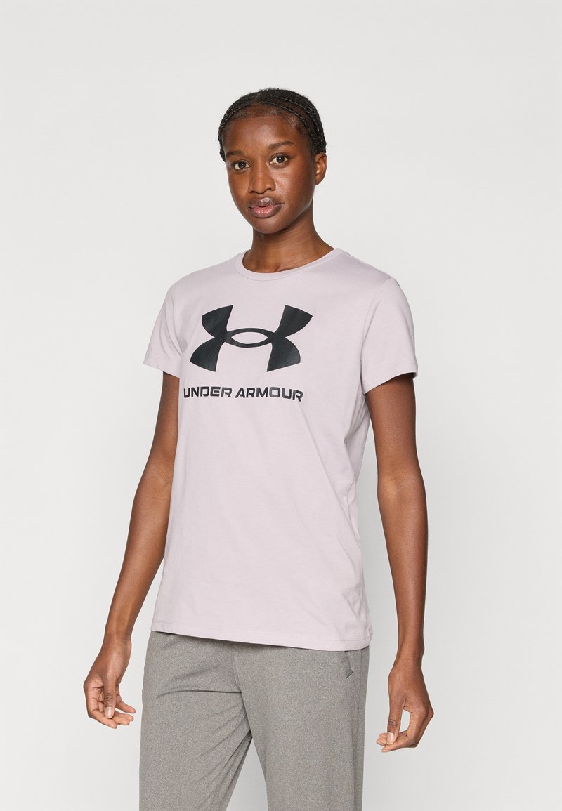 Femme debout portant un t-shirt Under Armour rose clair avec un logo noir et un pantalon gris sur un fond clair uni.