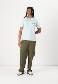 Lys blå T-skjorte med korte ermer og rød "Billabong" logo, sammen med olivengrønne cargo-bukser. Modellen har på seg hvite joggesko. Enkel bakgrunn.