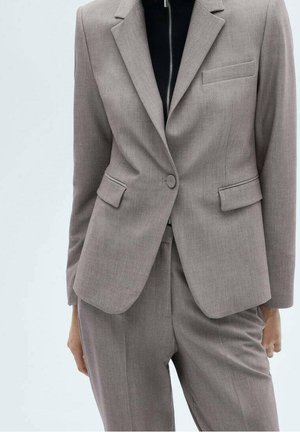 Kobieta w dopasowanym jasnoszarym garniturze z marynarką na jeden guzik i dopasowanymi spodniami, zestawiona z czarnym topem na zamek pod spodem.