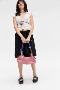 Woman wearing white ruffled blouse and black skirt, holding pink handbag with blue patterned handle, wearing black buckle shoes.