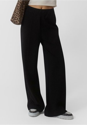 Crne široke hlače izrađene od mekanog materijala, s visokim strukom i elegantnim, minimalističkim dizajnom. Uparene sa sivim tenisicama.