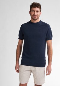 Navy modrý pletený svetr s kr átkým rukávem a kulatým výstřihem, s ribbovaným lemem a manžetami. V kombinaci s béžovými proužkovanými šortkami.