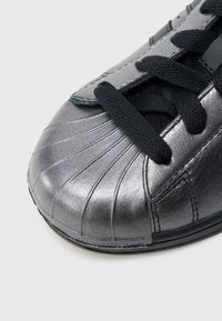 Μεταλλικό γκρι αθλητικό παπούτσι με στρογγυλεμένο μύτη, υφή στο πάνω μέρος, μαύρα κορδόνια και αντ�ιθετικές μαύρες λεπτομέρειες. Διαθέτει κάθετες ριβδώσεις.