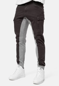 Cargo-broeken met een gespleten ontwerp in donkerbruin en lichtgrijze materialen, met ritszakken en elastische boorden. Draagt met witte sneakers.