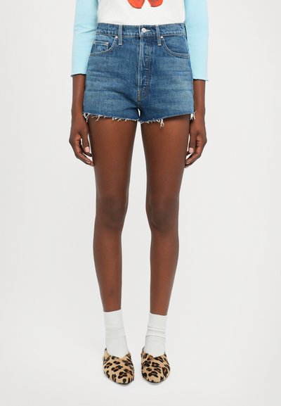 Benen in hooggetailleerde, versleten denim shorts, witte sokken en leuoenprint ballerina's tegen een eenvoudige witte achtergrond.