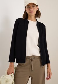 Veste zippée noire avec ourlet côtelé, portée par-dessus un haut beige clair et un pantalon khaki à cordon de serrage. Accentuée par une casquette crème.