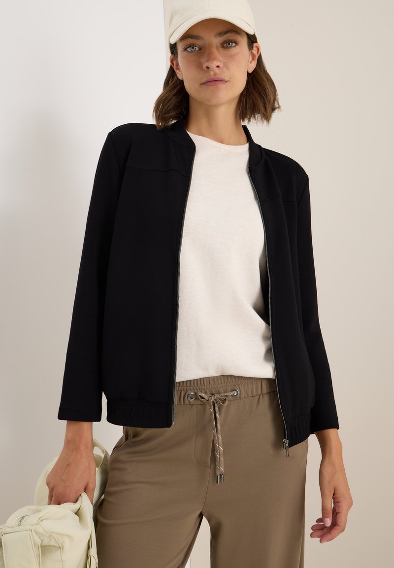 Veste zippée noire avec ourlet côtelé, portée par-dessus un haut beige clair et un pantalon khaki à cordon de serrage. Accentuée par une casquette crème.