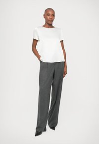 Γυναίκα με κοντά μαλλιά στέκεται με τα χέρια στις τσέπες, φορώντας λευκό κοντομάνικο μπλουζάκι, γκρι φαρδιά παντελόνια και μαύρα παπούτσια με αιχμηρή μύτη.