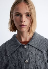 Grauer Strickcardigan mit großem Kragen, silbernen Knopfakzenten und Rippstruktur, getragen über einem rostfarbenen Oberteil.