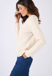 Ivoor gebreide trui met een v-hals, ribbelstructuur en kabeldetails; gecombineerd met donkerblauwe jeans voor contrast.