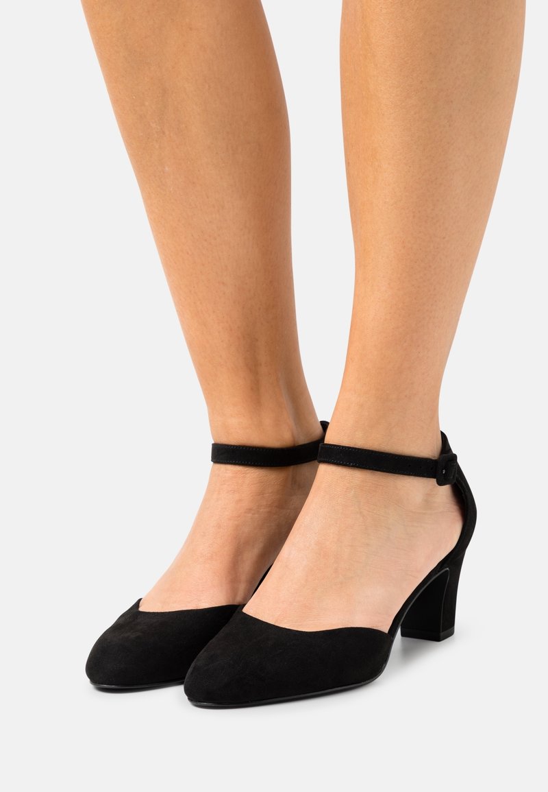 Jambes portant des escarpins noirs en daim à bout fermé avec des brides à la cheville et des talons blocs, se tenant devant un fond clair uni.